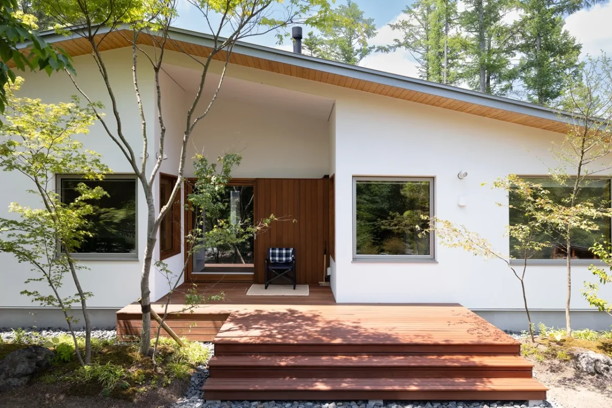 木々に囲まれた白い外壁の住宅と、庭に続くデッキ