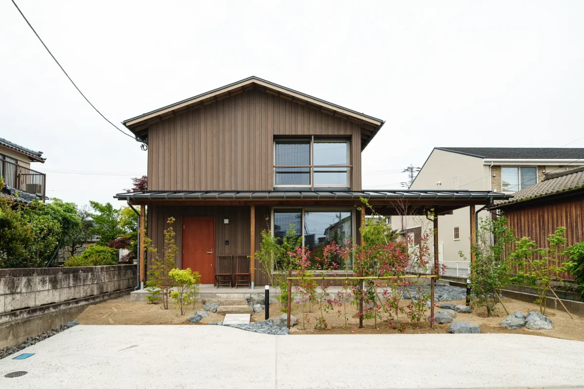 茶色い縦張りサイディングの2階建て住宅の外観。赤い玄関ドアと大きな窓が特徴で、前庭には砂利と植栽がある