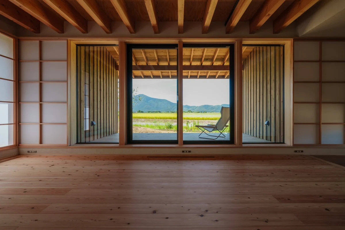 木造の天井と床を持つ室内から、引き戸越しにデッキと田園風景、山々を眺める景観