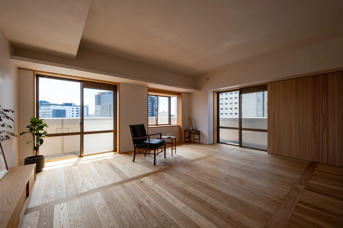 木製フレームの大きな窓から都市風景を望む、明るいリビング空間。浅い色の木製フローリング、ベージュ色の壁、モダンな黒いチェア、観葉植物が配置されている。